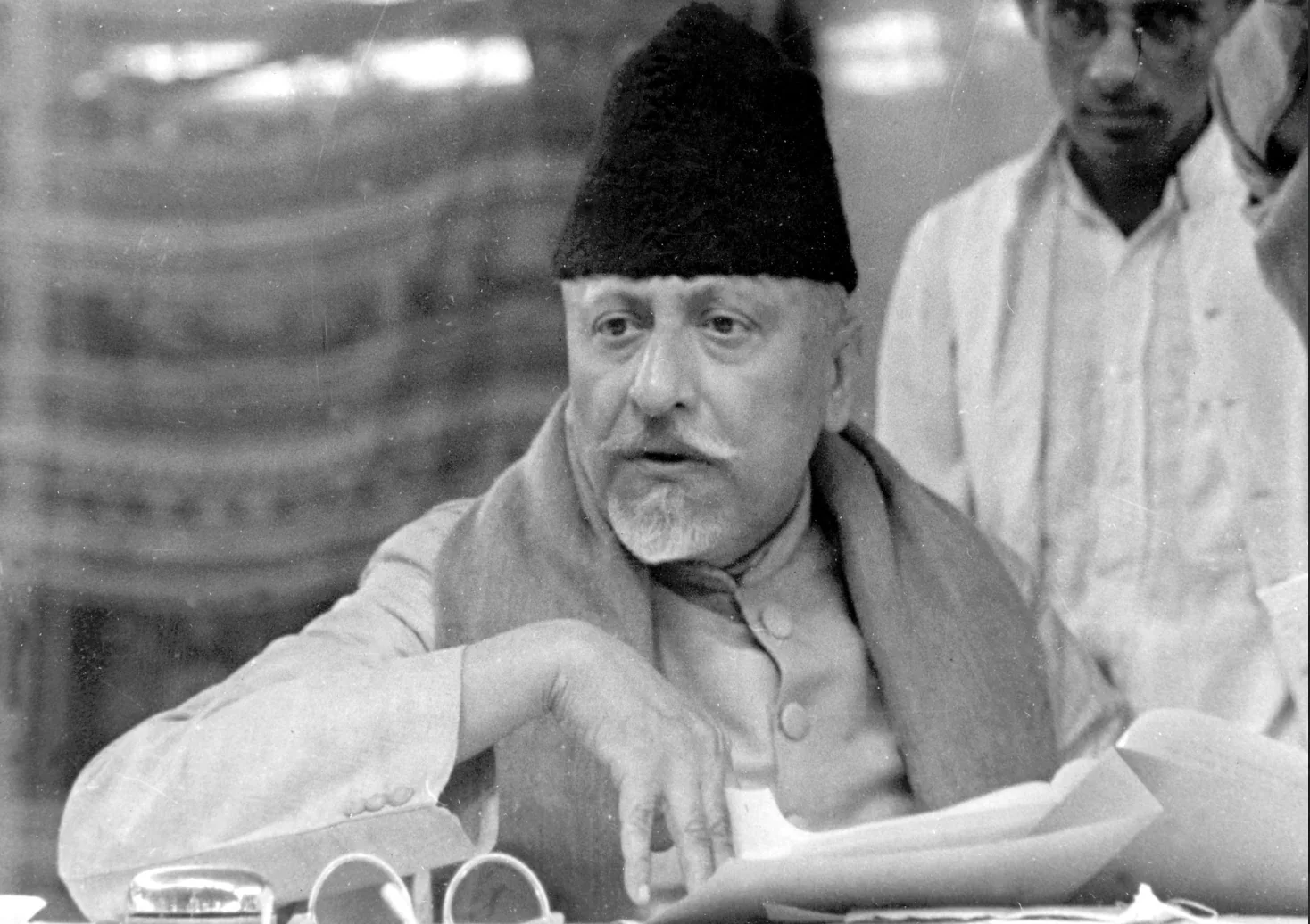 Maulana Abul Kalam Azad seated at a table covered with papers and spectacles, speaking, with others standing behind him.
